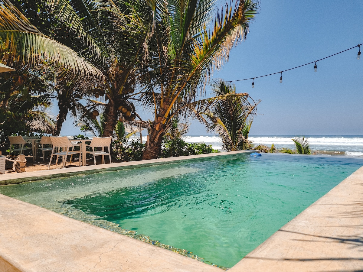 An infinity pool is seen with crystal-clear water, surrounded by lush palm trees. White outdoor dining tables and chairs are positioned nearby, providing a view of the ocean waves in the background. Soft sunlight enhances the inviting atmosphere.