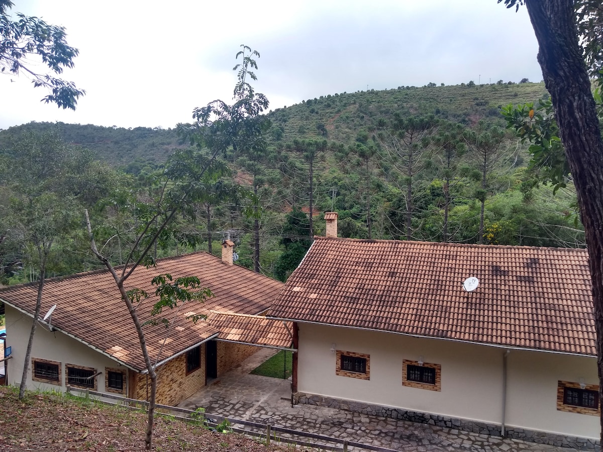 The exterior of a spacious house is shown, featuring a tiled roof and stone accents. A well-maintained path leads to the entrance. Surrounding greenery includes trees and hills, creating a serene natural environment.
