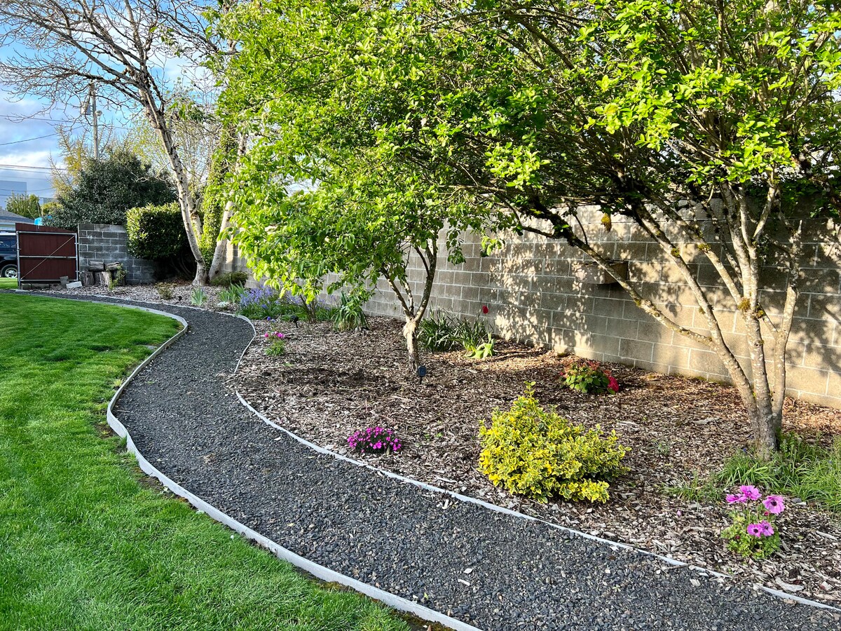 A winding path lined with gravel meanders through a landscaped area, featuring lush green grass and flowering plants. Trees provide shade along the pathway, while a stone wall offers privacy in the background. Fresh blooms add color to the serene outdoor space.