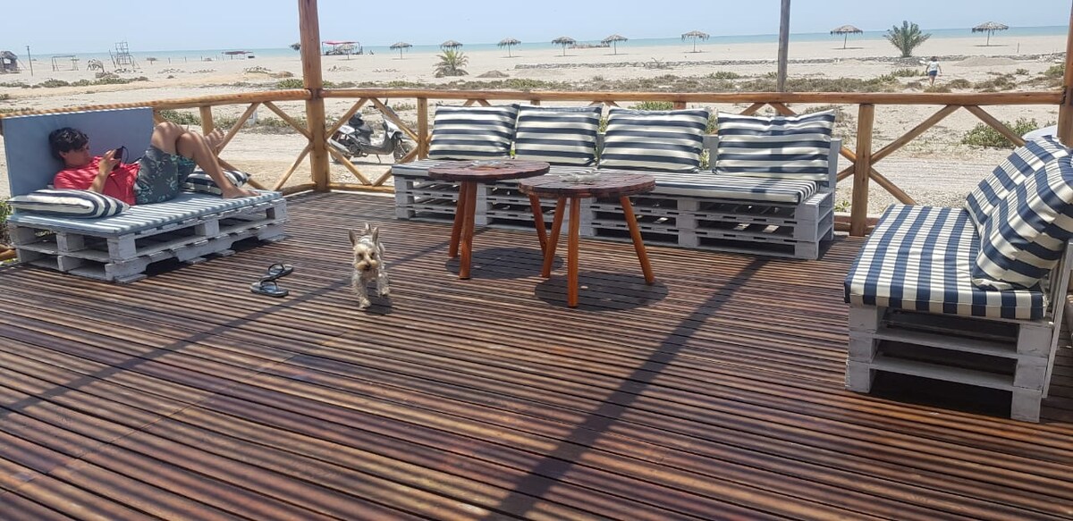 A spacious terrace features several seating areas arranged with striped cushions atop wooden pallets. A small wooden table sits in the center. The beach, visible in the background, is lined with umbrellas and palm trees, enhancing the serene seaside atmosphere.
