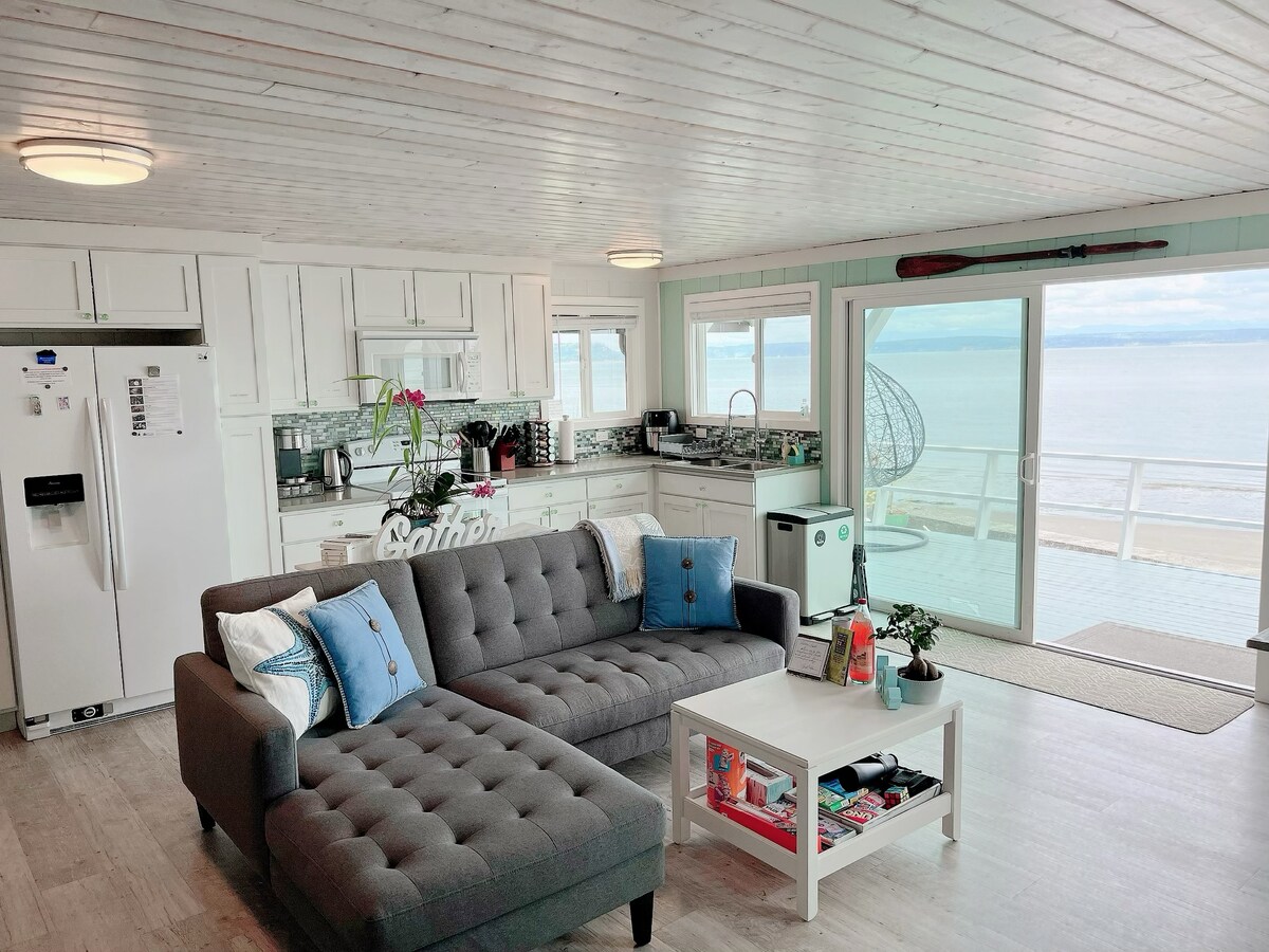 A modern open-concept space showcases a comfortable gray sectional sofa paired with a white coffee table, positioned to take in ocean views. The well-equipped kitchen features white cabinetry and stainless steel appliances, with a glass door leading to an outdoor deck overlooking the water.
