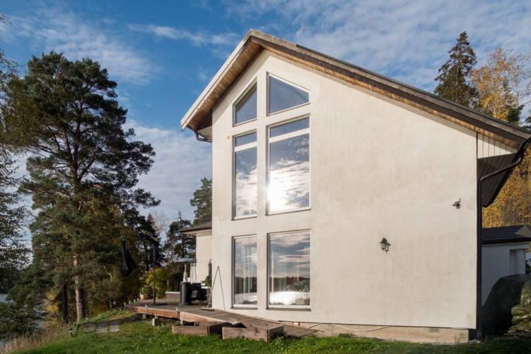 An exterior view of the house is showcased, featuring large, modern windows that reflect the sky. The structure emphasizes a contemporary design with a sloped roof and wooden deck, surrounded by trees that enhance the natural setting by the lake.