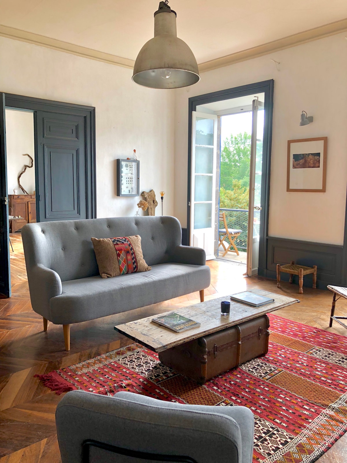 A seating area is presented with a light gray sofa featuring decorative cushions. A rustic wooden coffee table rests on a colorful area rug. Large doors lead to a balcony, inviting natural light into the room, with framed artwork adorning the walls.