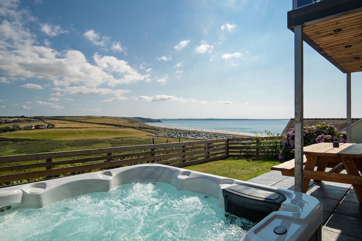 A luxurious hot tub is positioned on a terrace, offering expansive views of the rolling hills and sea. The clear blue sky and fluffy clouds enhance the scenic backdrop. A picnic table sits nearby, inviting outdoor relaxation amid natural beauty.