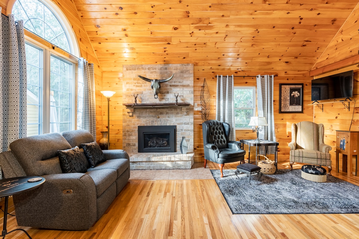 A spacious living area features warm wood-paneled walls and a cathedral ceiling. A gas fireplace is set against a stone backdrop, flanked by two comfortable armchairs and a sofa. Natural light filters through a large arched window, enhancing the inviting atmosphere.