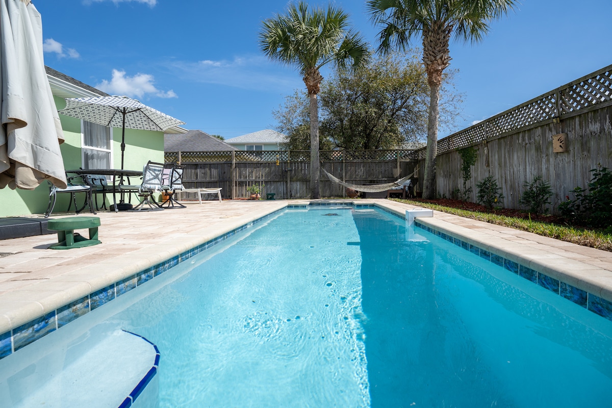 A private backyard features a heated swimming pool surrounded by tiled patio. Lush palm trees rise above the pool area. A hammock rests in the corner, inviting relaxation. Outdoor furniture is visible, along with a sun umbrella providing shade.