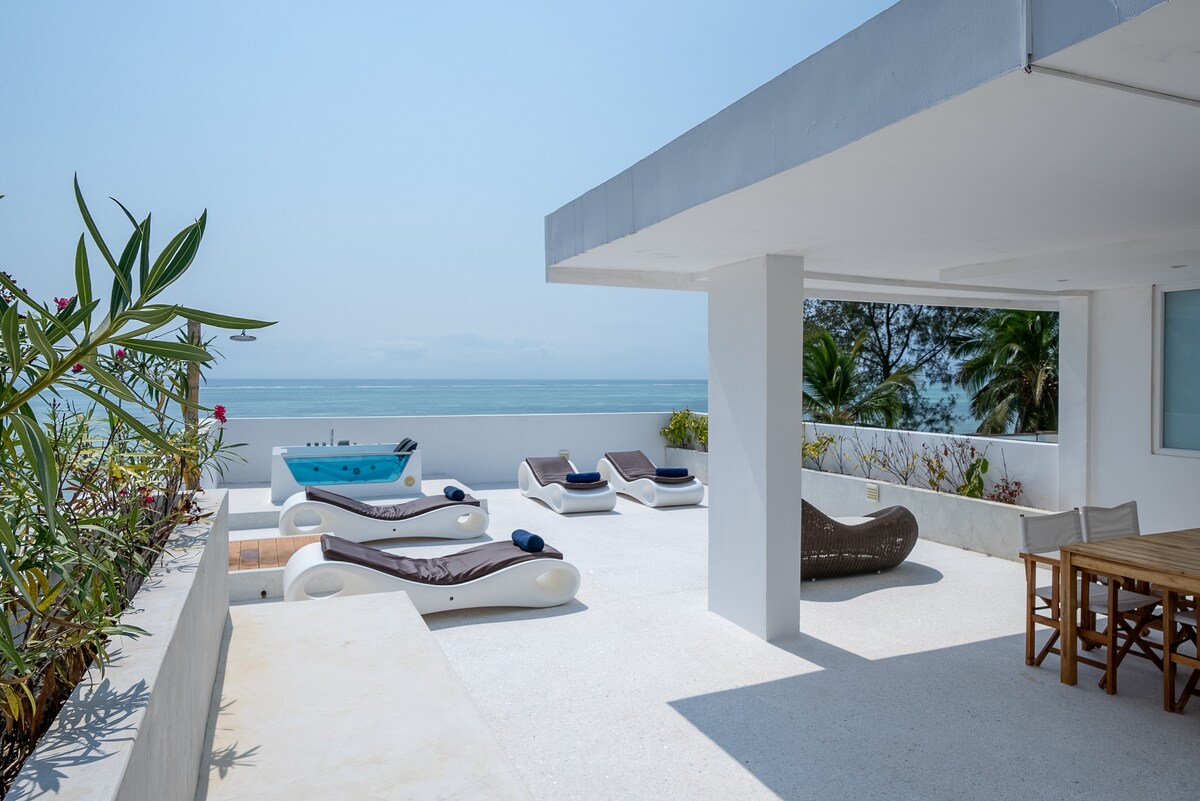 A spacious rooftop terrace features a dining table, multiple sun loungers, and a hot tub, all with unobstructed ocean views. Lush greenery lines the space, adding a touch of nature to the modern setting. The bright atmosphere is complemented by ample sunlight.