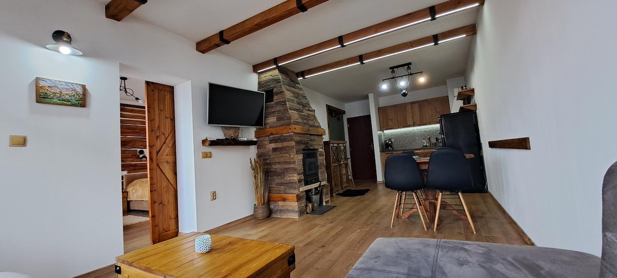 An open-plan living area features wooden beams overhead and a stone fireplace as the focal point. A comfortable seating arrangement is complemented by a wooden coffee table. Nearby, a kitchen is visible, showcasing modern appliances and warm wooden cabinetry.