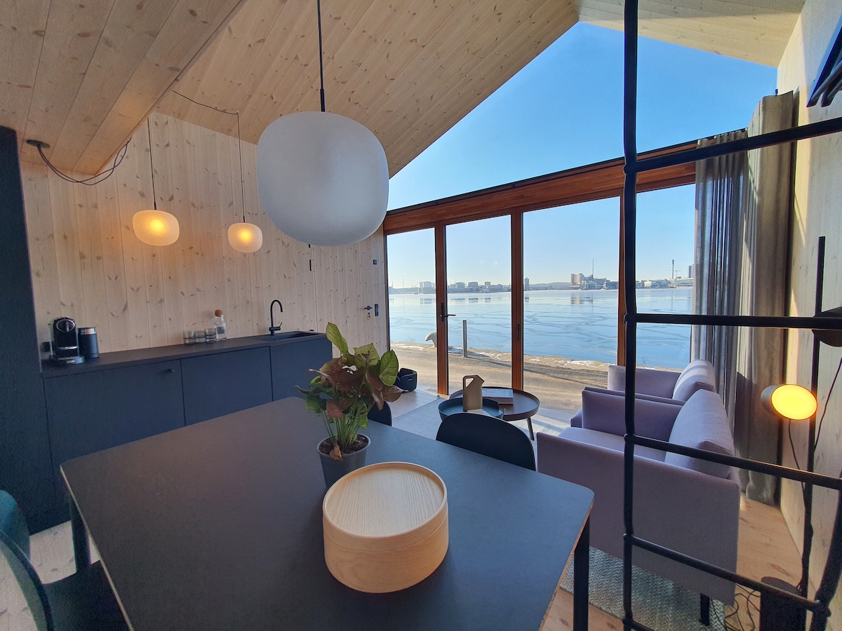 An open living area features a modern kitchen with a black countertop and a dining table. Large glass doors provide access to picturesque views of the harbor and city. A cozy seating space with a light sofa and a stylish pendant light complements the wooden ceiling.