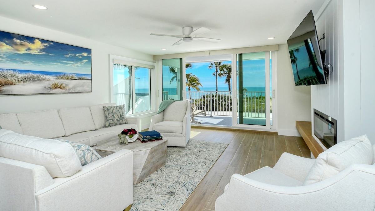 The living area features a light-colored sectional sofa and armchairs arranged around a decorative rug. Large windows provide unobstructed views of the beach and gulf, complemented by a modern entertainment unit and artwork that reflects the coastal setting.
