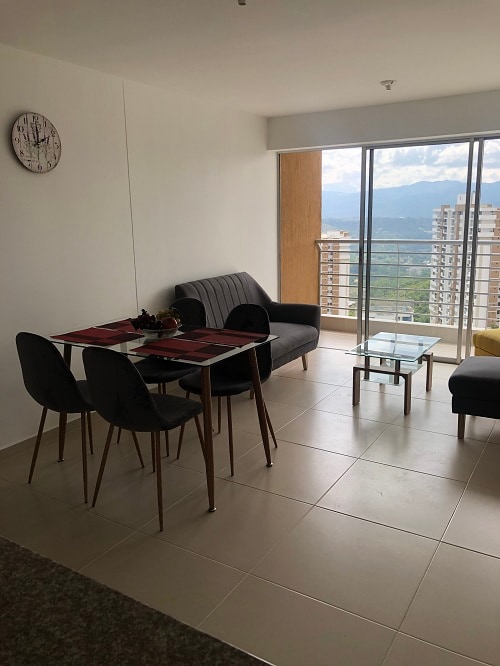 A modern living area is displayed with a dining table set for four and a comfortable gray sofa. Large glass doors lead to a balcony, offering views of the surrounding mountains. A clock hangs on the wall, enhancing the minimalist décor.