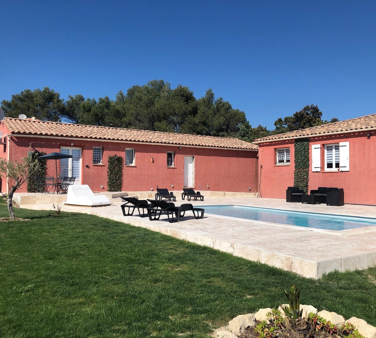 A modern, single-story residence features a warm pink exterior and a tiled roof. A spacious pool framed by sun loungers is surrounded by green grass and a variety of shrubs, with trees visible in the background under a clear blue sky.