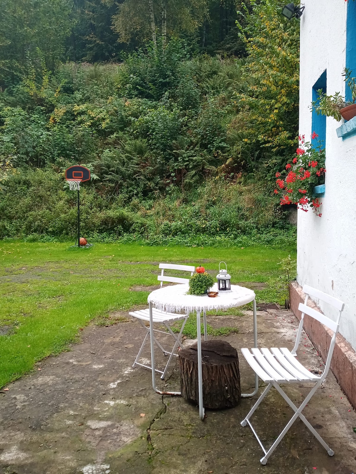 An outdoor seating area features a round table surrounded by two folding chairs. A lantern and a potted plant add a touch of charm. In the background, a basketball hoop stands among greenery, with a well-maintained lawn extending around the space.