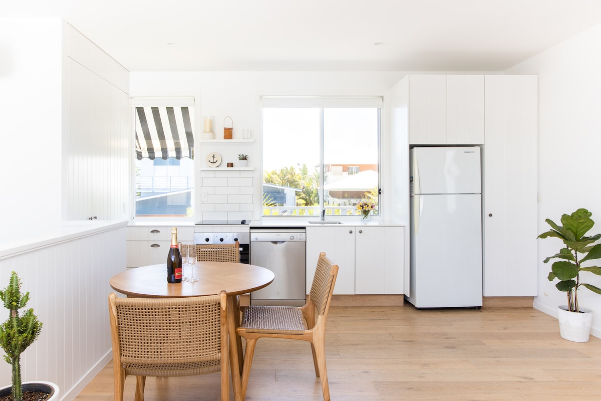 A modern kitchen and dining area is displayed, featuring a round wooden table with two wicker chairs. A large window allows natural light to fill the space, with a view of greenery outside. Stainless steel appliances, including a dishwasher, are visible in the sleek cabinetry.