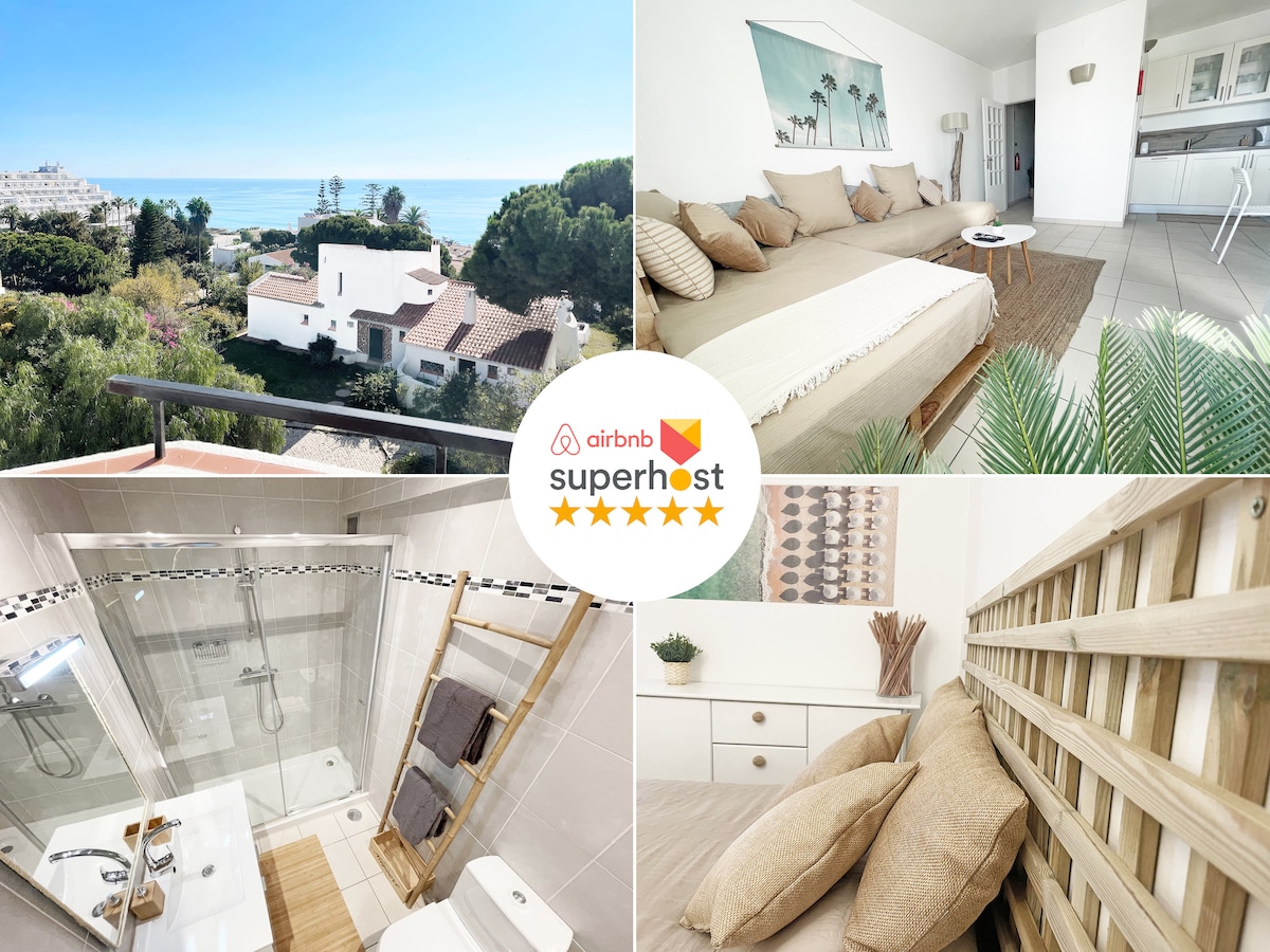 A sunlit, open living space reveals a spacious bed adorned with neutral linens, complemented by decorative pillows. A scenic view of the ocean and welcoming greenery is visible through the glass doors. The bathroom features modern fixtures, with a stylish ladder shelf adding character.
