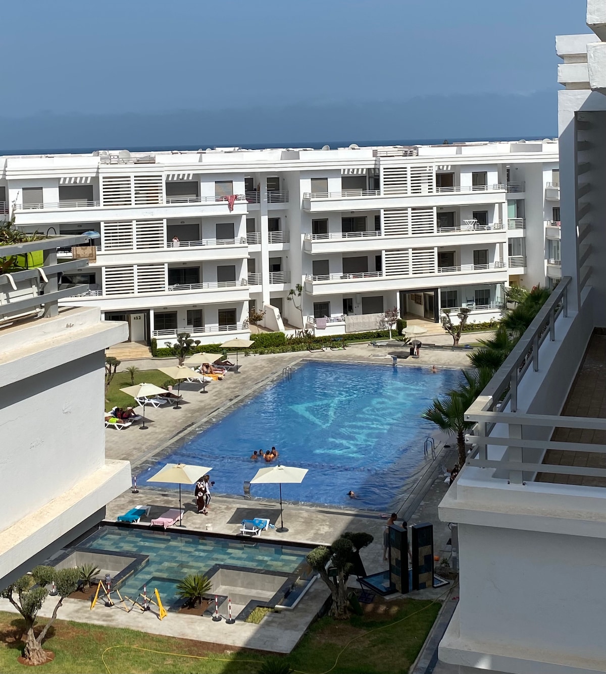 An aerial view captures a swimming pool surrounded by lounge chairs and umbrellas, with residents enjoying the water. The contemporary architecture of the surrounding buildings is visible, complemented by green landscaping and a glimpse of the ocean in the background.