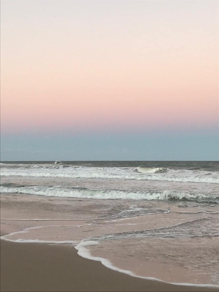 The image captures a serene beach scene, where gentle waves lap against the shore. A soft gradient of pastel hues transitions from pink to blue in the sky, reflecting the peacefulness of the coastal environment.
