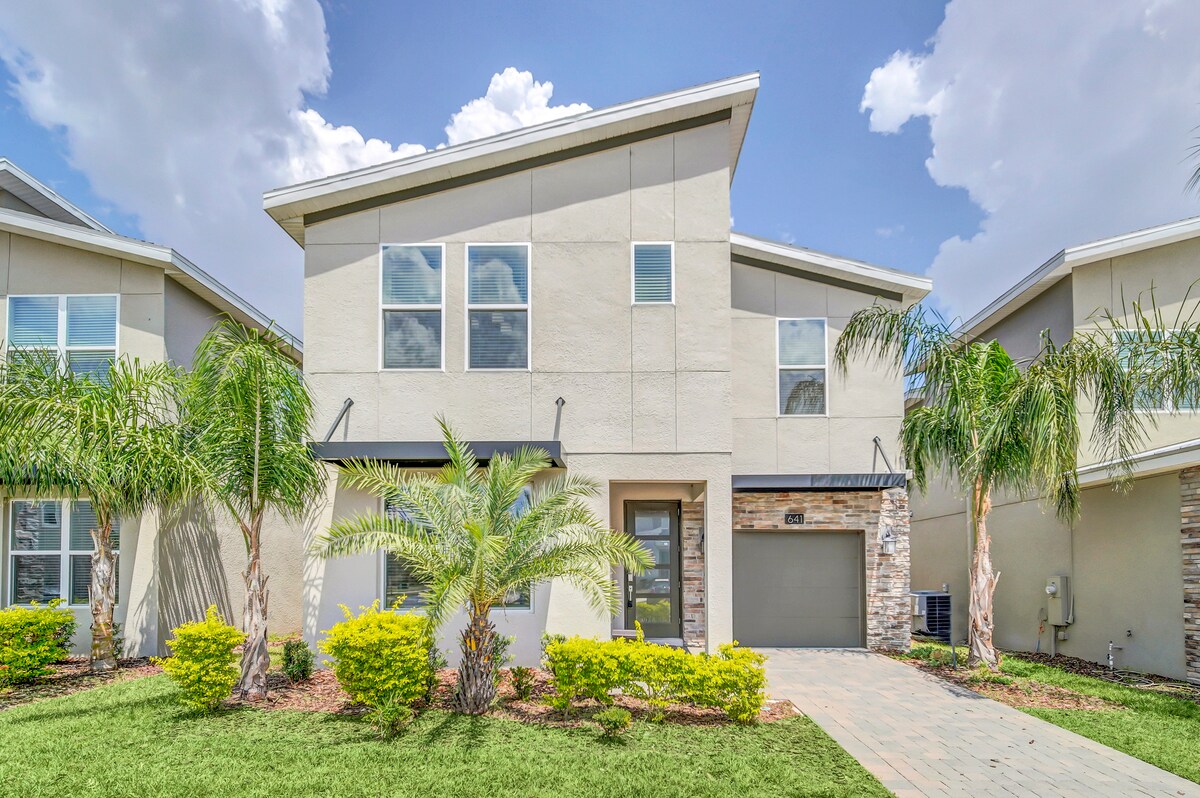 The exterior of the house features a modern two-story design, framed by lush palm trees and well-maintained landscaping. A paved pathway leads to the entrance, while large windows allow ample natural light to illuminate the interior.