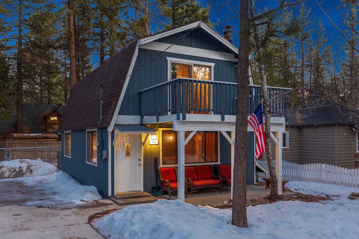 A charming gambrel-style cabin is framed by tall pine trees, featuring a wraparound porch with red seating. An American flag is displayed on the exterior, while snow blankets the ground. Warm light emanates from the windows, creating a welcoming evening scene.