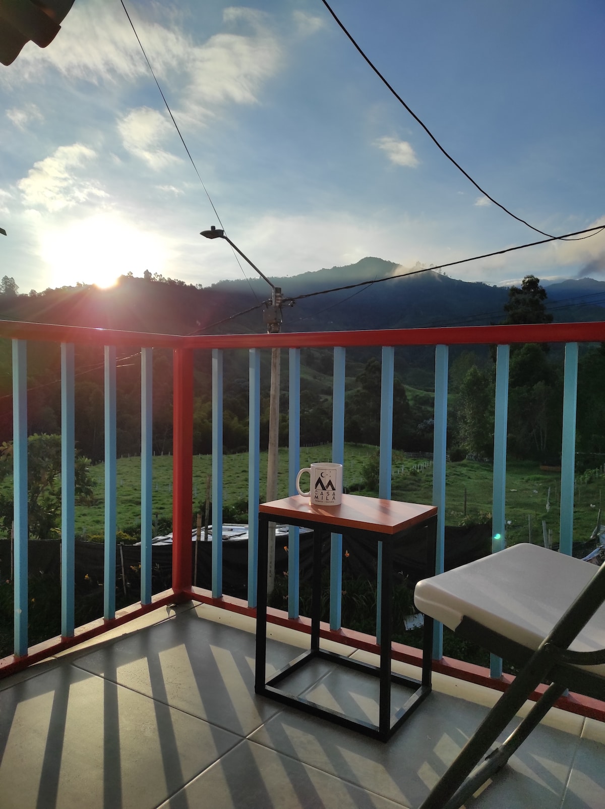 A small table with a cup sits on the balcony, framed by colorful railings. The sun rises behind distant mountains, illuminating the green landscape below. Clear skies suggest a serene morning atmosphere.