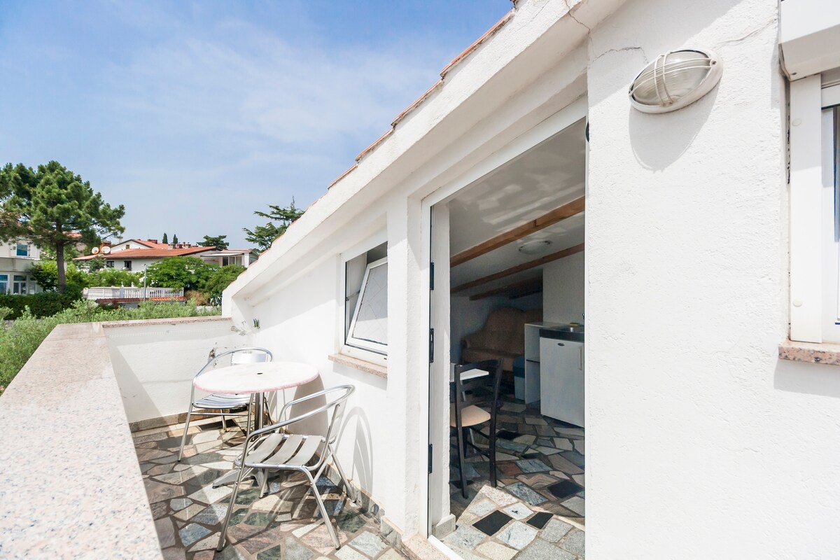 A small terrace is featured in front of the entrance, equipped with a round table and two chairs. The terrace overlooks a green space and is characterized by its stone-tiled surface. Natural light enhances the area, with a sloped ceiling visible in the background.