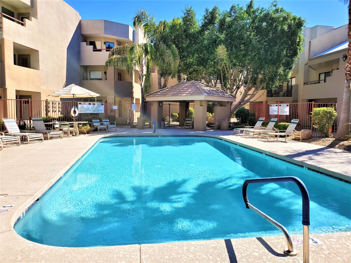 A clear view of a community pool surrounded by lounge chairs. The pool’s blue water reflects the sun, with palm trees providing shade nearby. A gazebo with seating is present in the background, indicating a comfortable outdoor space for relaxation.