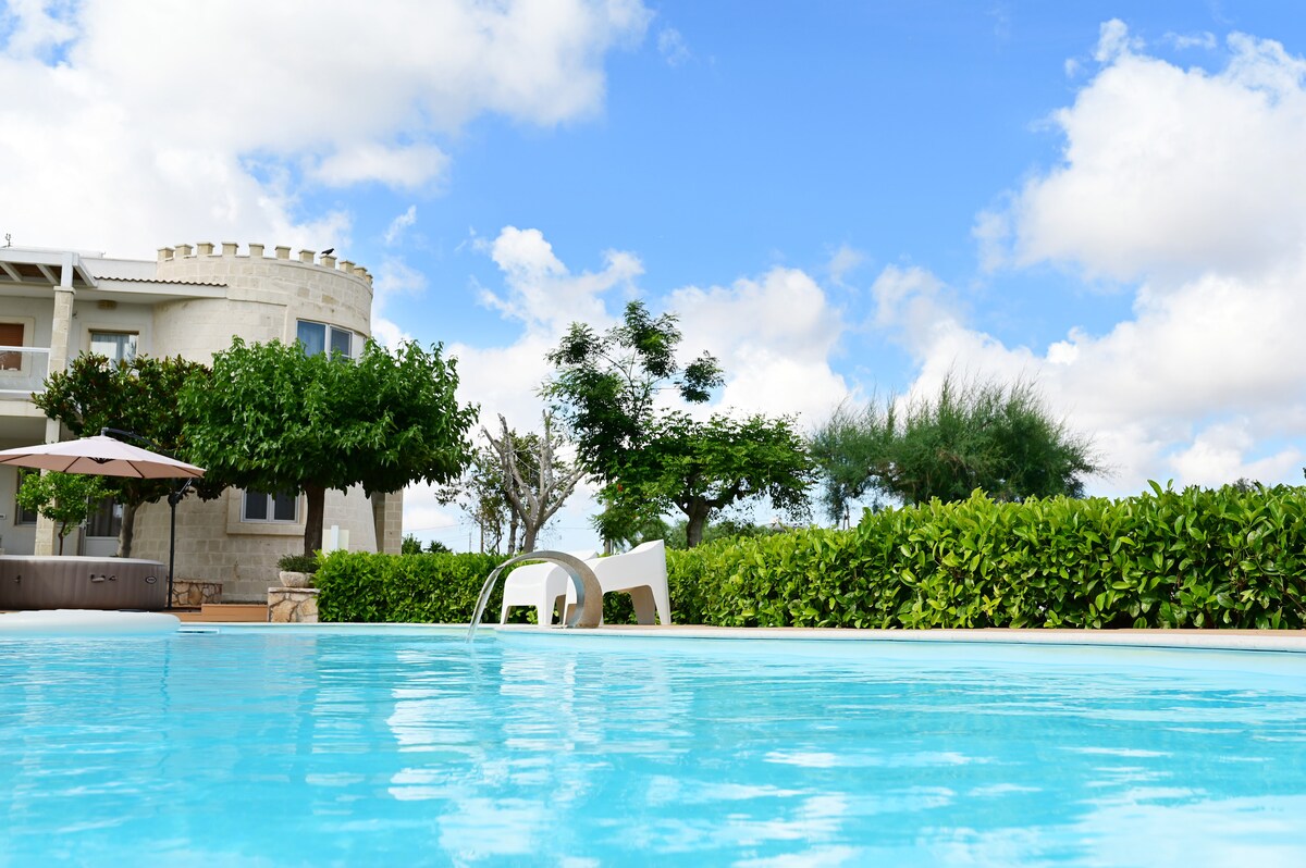 A clear swimming pool reflects the bright blue sky, surrounded by neatly trimmed hedges and green trees. The structure in the background features a castle-like tower and outdoor seating areas under umbrellas, integrating with the serene garden setting.