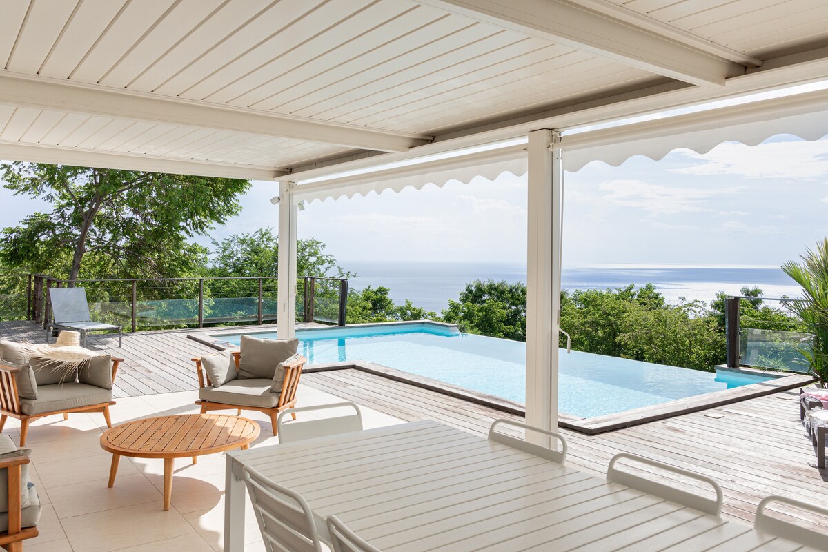 An outdoor terrace features a wooden dining table surrounded by white chairs, overlooking a serene infinity pool. Comfortable lounge chairs are arranged nearby, creating a relaxed seating area. Lush greenery frames the view, which extends towards the horizon and the ocean.