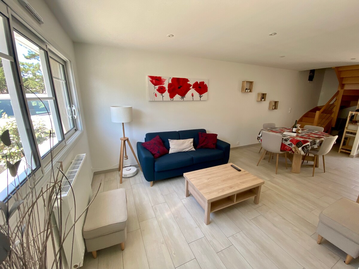 A bright living area features a navy blue sofa adorned with red pillows, complemented by a wooden coffee table. A lamp stands beside the sofa, and a dining area with a checkered tablecloth is visible nearby. Light streams in through large windows, enhancing the spacious feel.