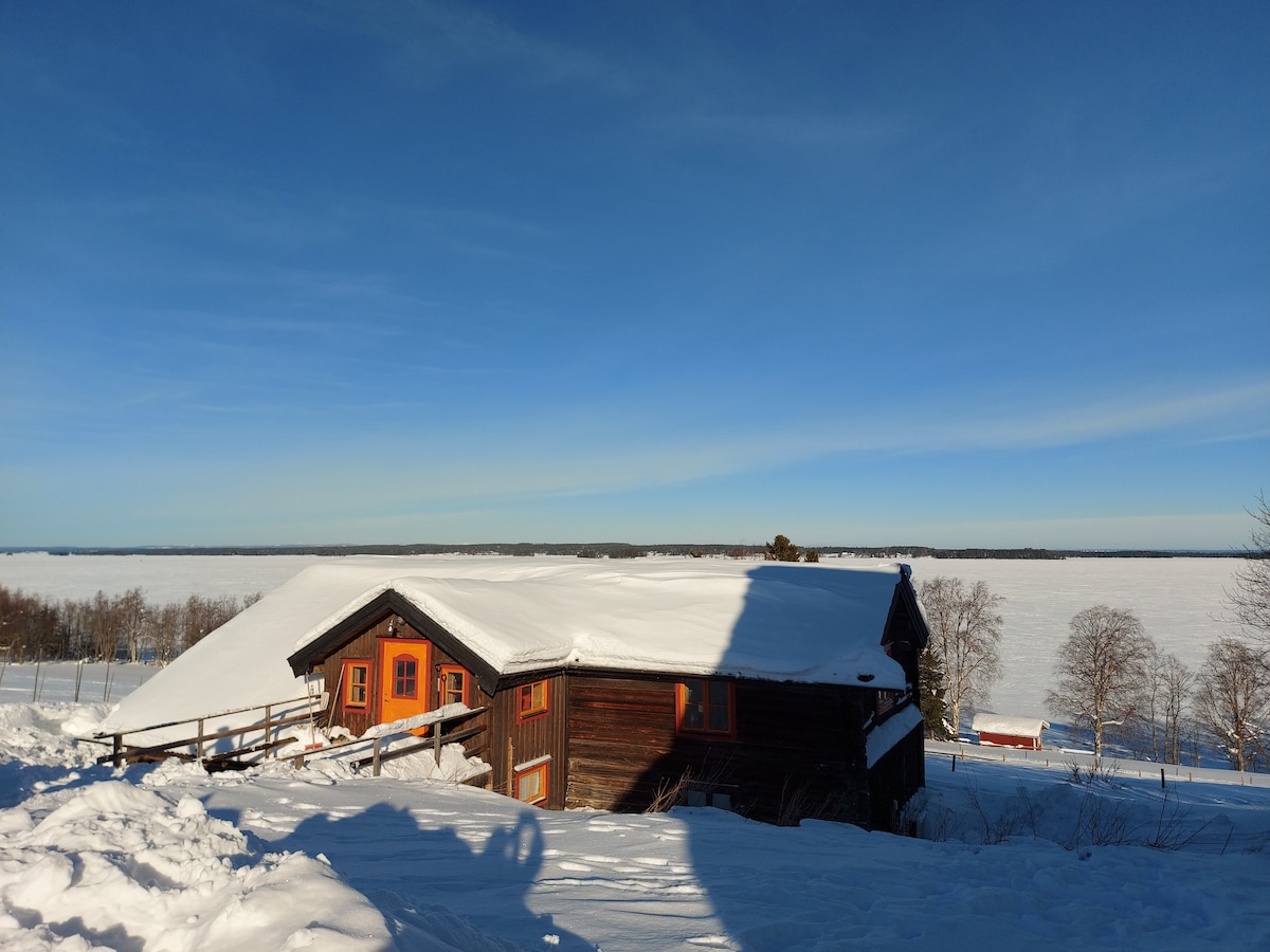 The rustic cabin is partially covered in snow, showcasing its wooden structure and bright orange window frames. The frozen Storsjön lake stretches out in the background under a clear blue sky, with nearby trees outlined by snow.