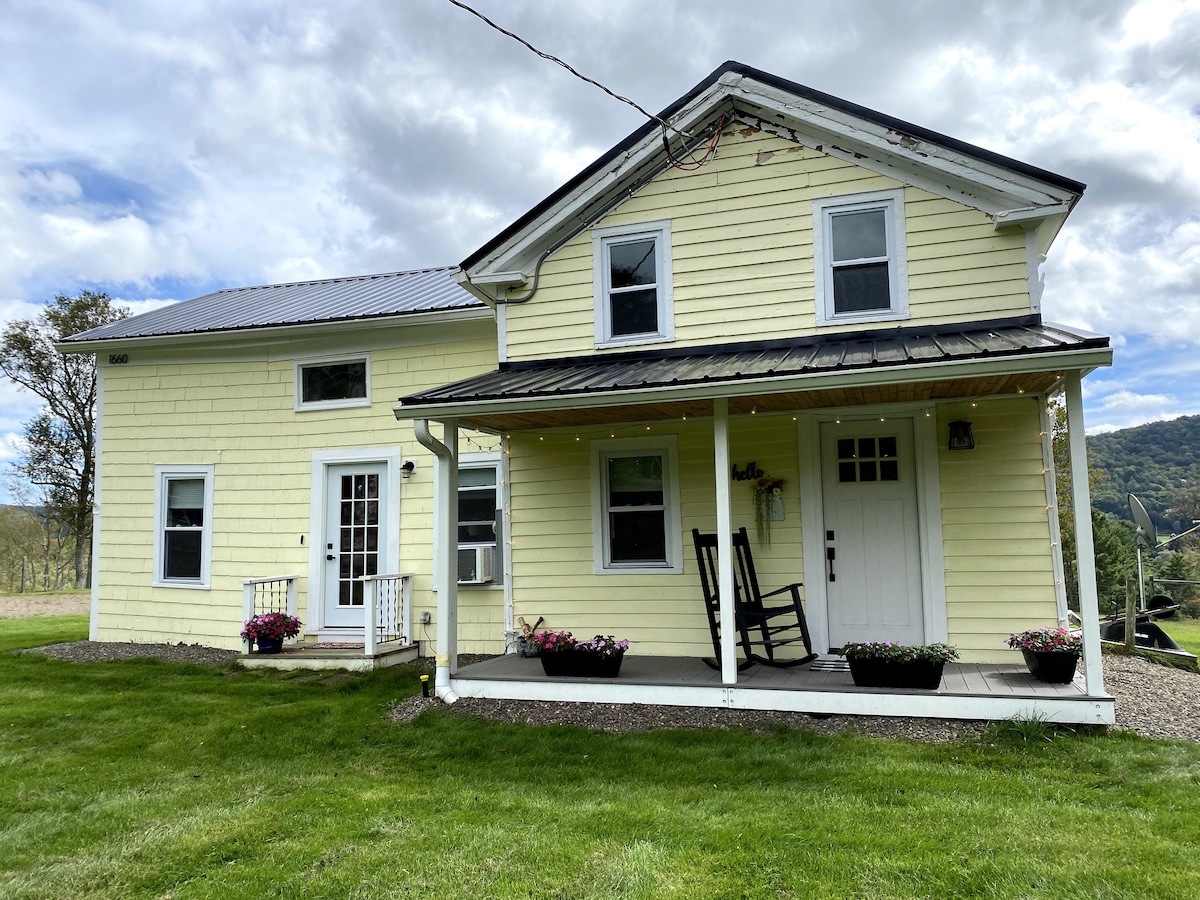 The charming farmhouse is presented in a soft yellow hue, featuring a covered porch with two rocking chairs. Flower pots with colorful blooms are placed on either side of the porch, and large windows allow natural light to flood the interior.