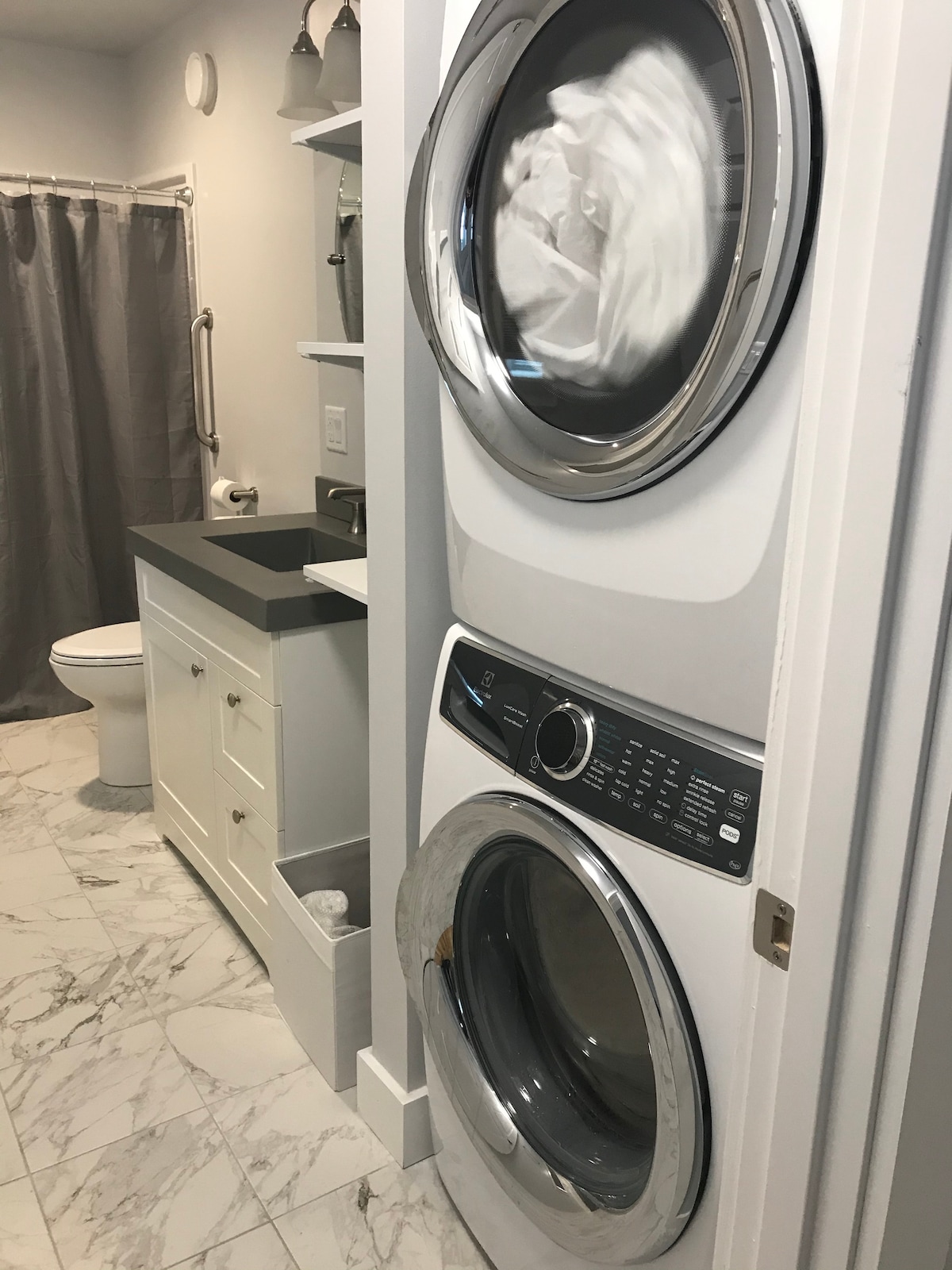 A laundry area is located adjacent to a bathroom, featuring a stacked washer and dryer with a sleek design. Light marble flooring contrasts with the white cabinetry, while a gray shower curtain provides a touch of color. The bathroom includes a toilet and a dark countertop.