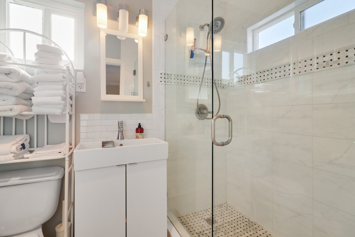 A modern bathroom features a minimalist design with a clear glass shower enclosure. Fresh white towels are neatly stacked on a rack beside a compact vanity with storage. Natural light enters through multiple windows, illuminating the neutral tones of the tiled walls and floor.