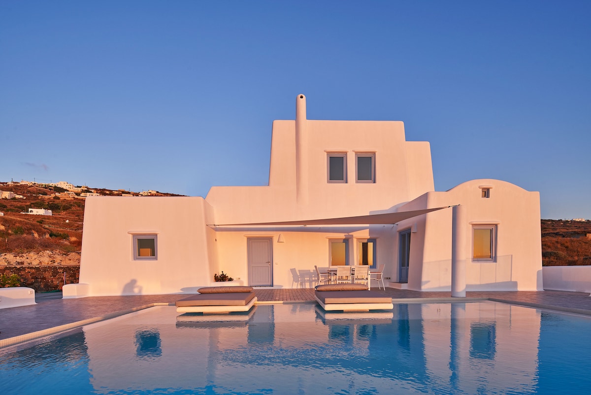 The villa's exterior is characterized by a minimalist white facade, with large windows reflecting the evening light. A spacious pool sits in the foreground, surrounded by a stone patio, while a shaded outdoor dining area is positioned nearby, inviting relaxation.