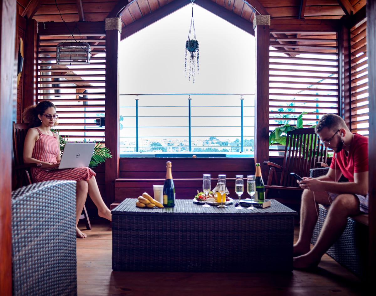 A covered balcony area is furnished with wicker seating and a coffee table. Various beverages and fruits are arranged on the table. The balcony overlooks a scenic view through slatted wooden panels, allowing natural light to filter into the space.