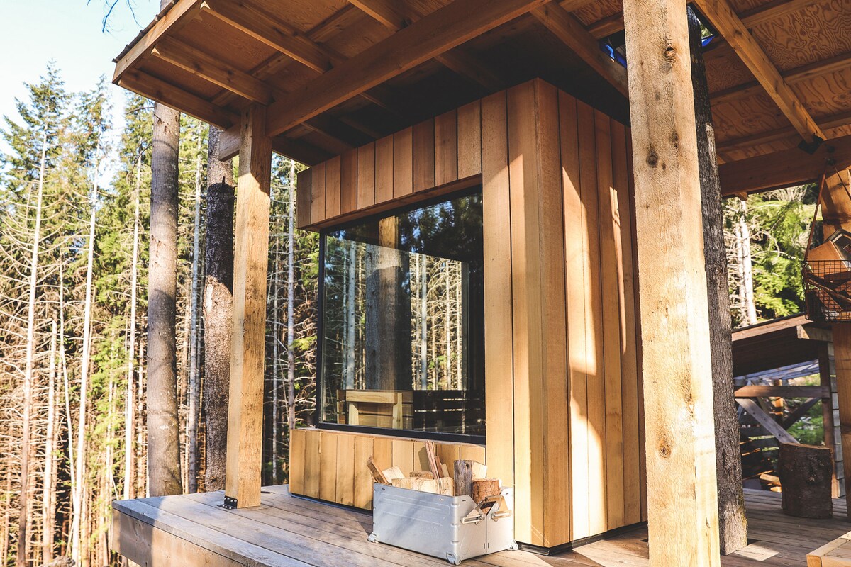 A wooden sauna structure blends with the natural surroundings, featuring large glass windows that offer views of the forest. A wood storage box is positioned outside, complementing the warm tone of the wood paneling and enhancing proximity to nature.