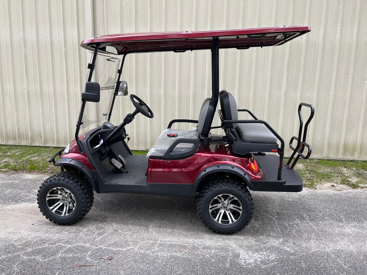 A red golf cart is positioned on a paved surface, displaying an open front with a steering wheel and two seats. The vehicle features large black tires and a canopy overhead, providing shade. The background consists of a plain, light-colored wall.