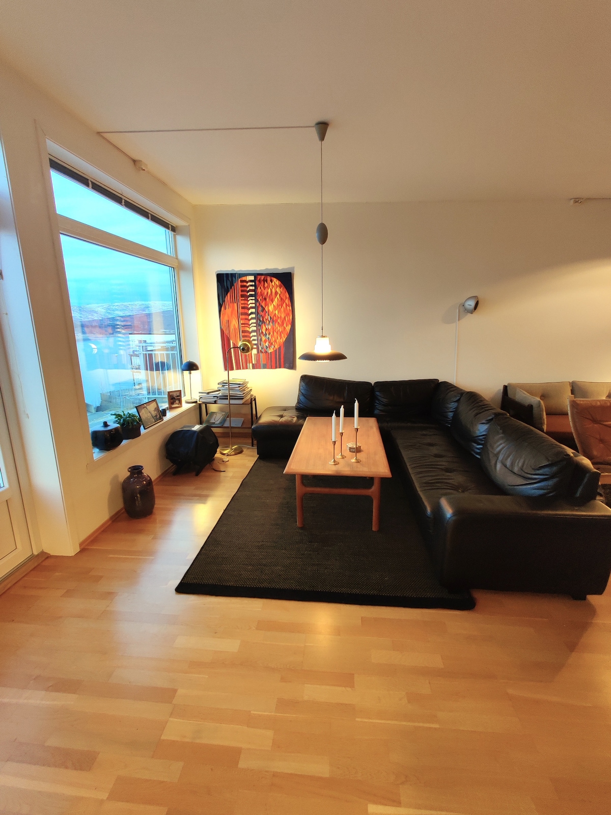 A spacious living area is highlighted by a large black sectional sofa facing a wooden coffee table. A modern pendant light hangs above the table, while large windows provide natural light and views. A colorful wall artwork adds a vibrant touch to the neutral decor.
