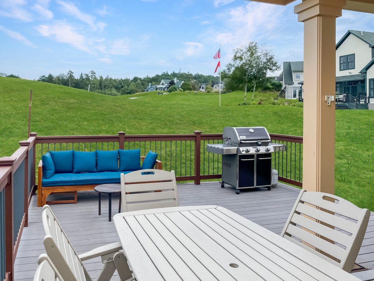 A spacious outdoor deck is featured, showcasing comfortable seating and a dining area. A grill is positioned nearby, and a verdant landscape extends beyond the deck. American and state flags can be seen gently waving in the breeze.