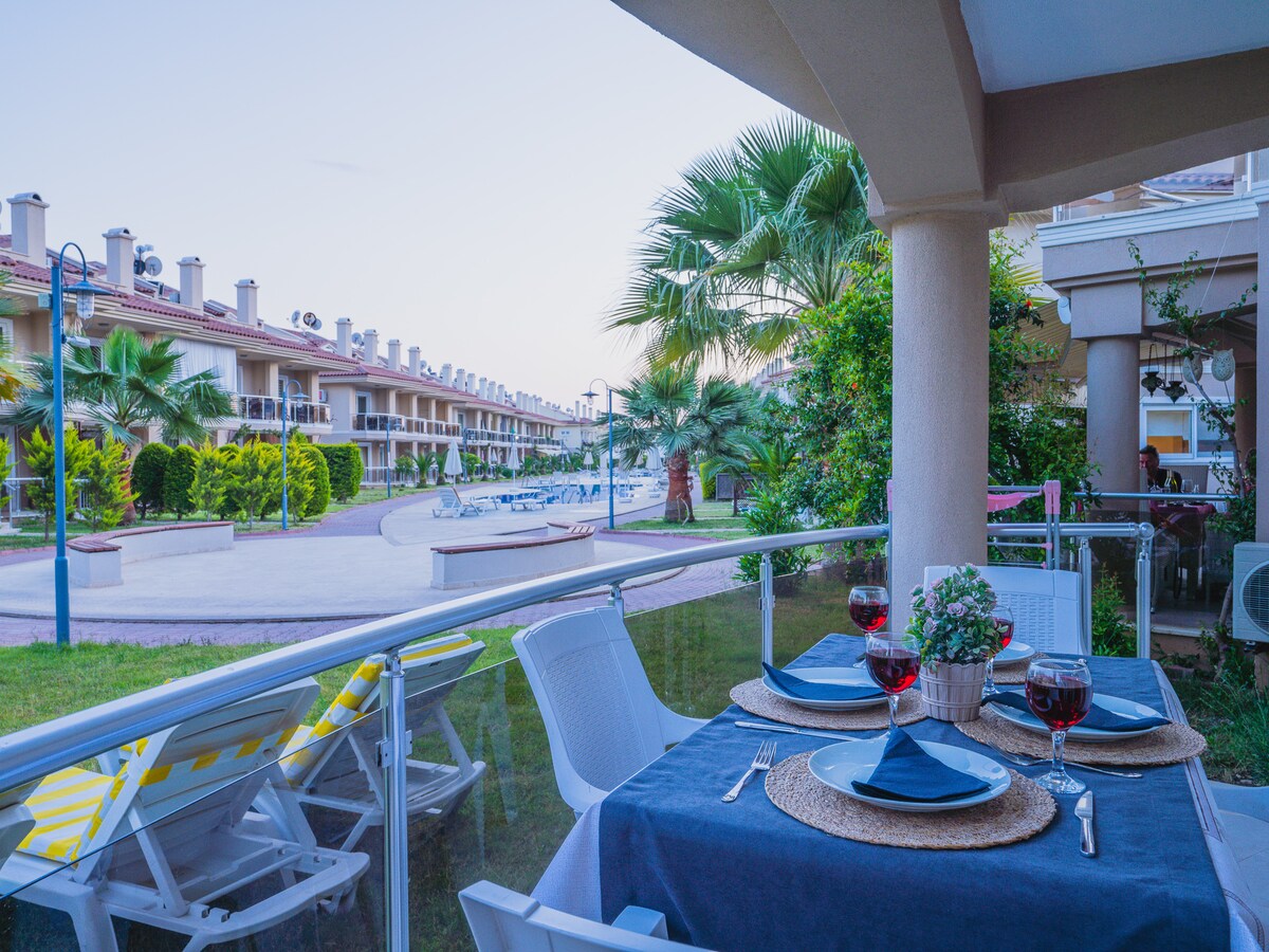 An outdoor dining area is presented, featuring a table set for two with glasses of red wine. Comfortable chairs are arranged around the table, and views of palm trees and a shared pool area are visible in the background. The ambiance conveys a relaxed atmosphere.
