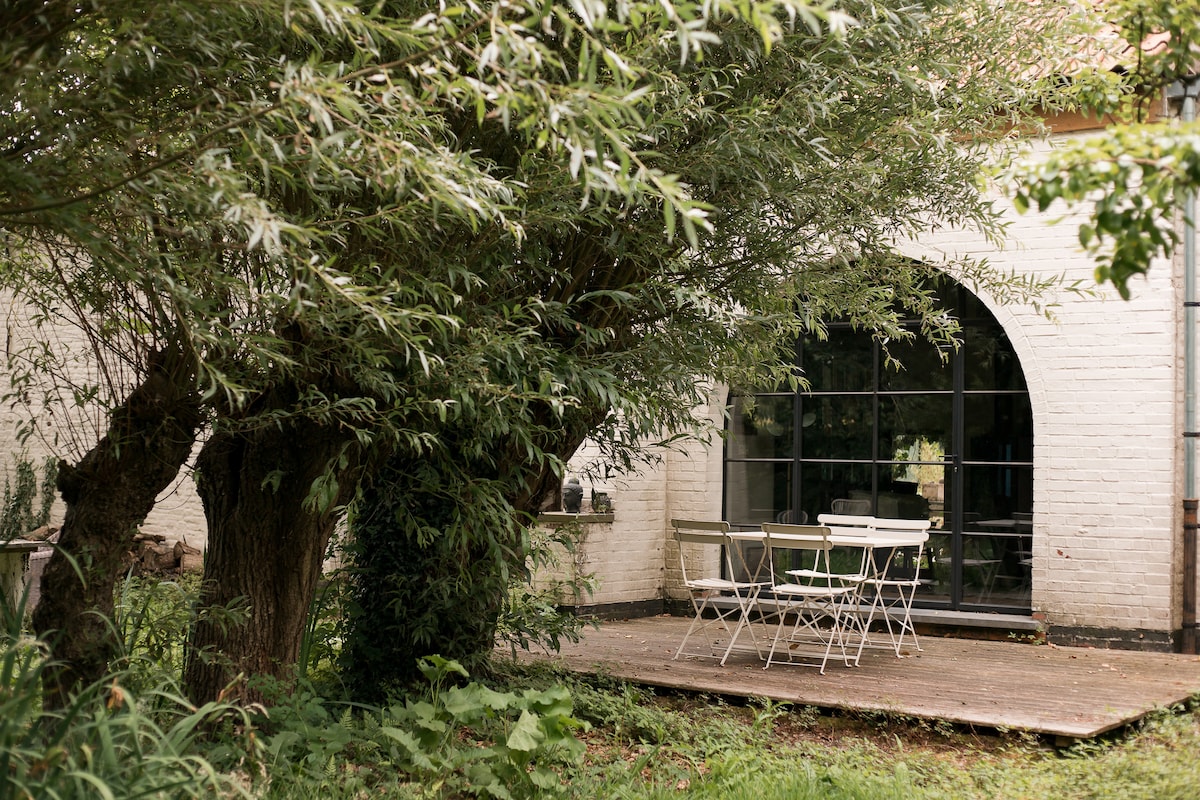 An outdoor seating area features a wooden deck surrounded by lush greenery. A round table is accompanied by four chairs, set against a backdrop of large arched windows with views of the garden.