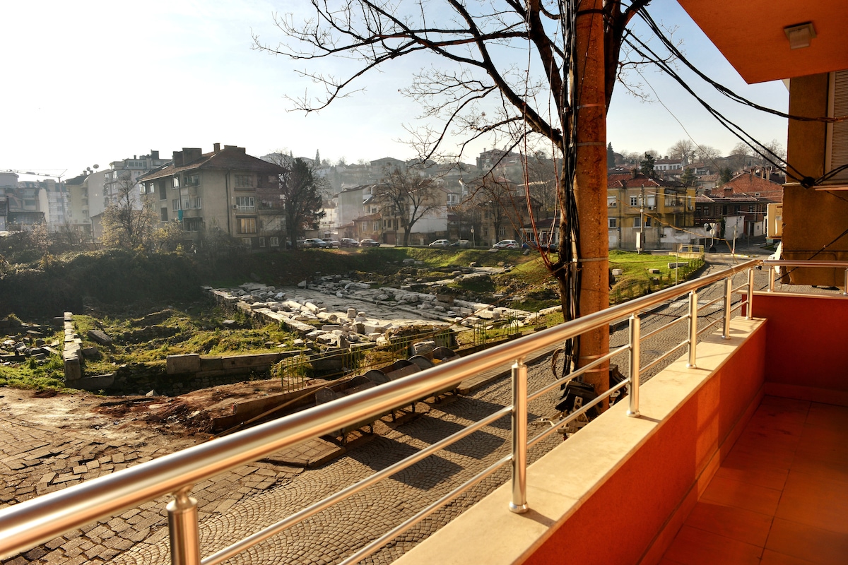A balcony view displays an open area featuring archaeological ruins, bordered by trees and buildings. The scene reveals a mix of natural and urban elements, with sunlight illuminating the landscape and railings providing depth to the perspective.