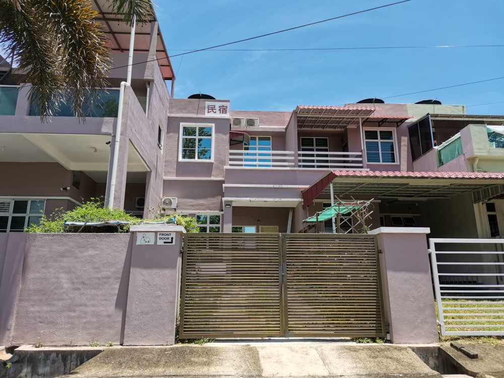 The exterior of the property is viewed, showcasing a modern two-story structure with a mix of pink and white facades. Air conditioning units are visible on the upper level. A decorative gate and surrounding greenery enhance the entrance, providing a welcoming atmosphere.