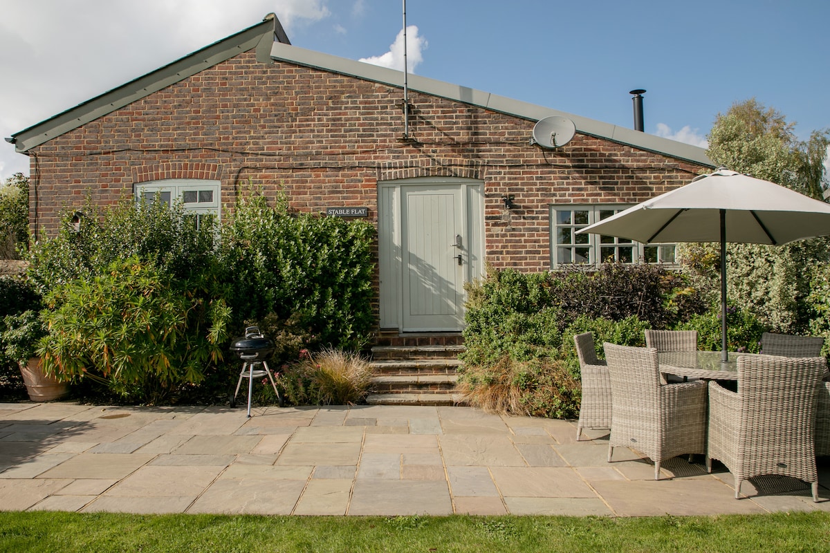 The exterior of the flat is shown, featuring a brick facade with a light green door and several windows. A patio area with a dining set and an umbrella invites outdoor meals. Lush greenery surrounds the space, creating a serene atmosphere.