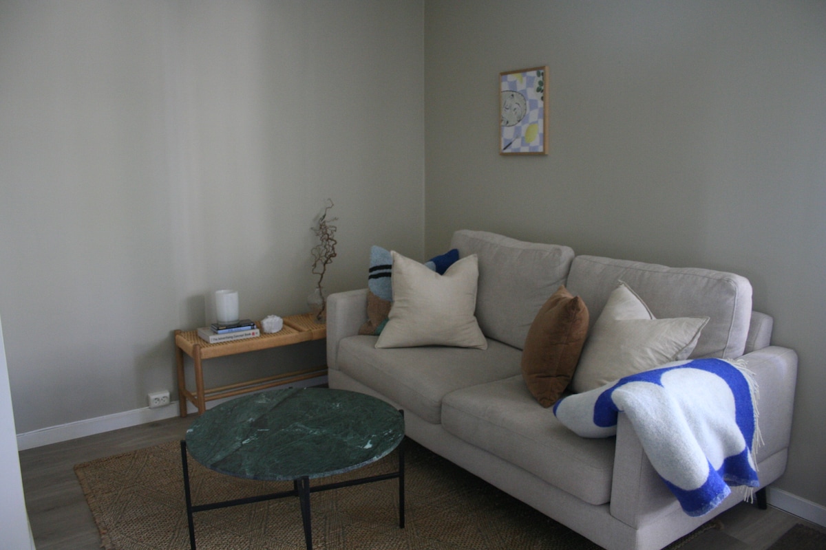 A cozy living area features a light grey sofa adorned with large cushions in neutral tones. A round green coffee table is positioned in the center, complemented by a woven rug beneath. A simple artwork hangs on the wall, adding a touch of character.