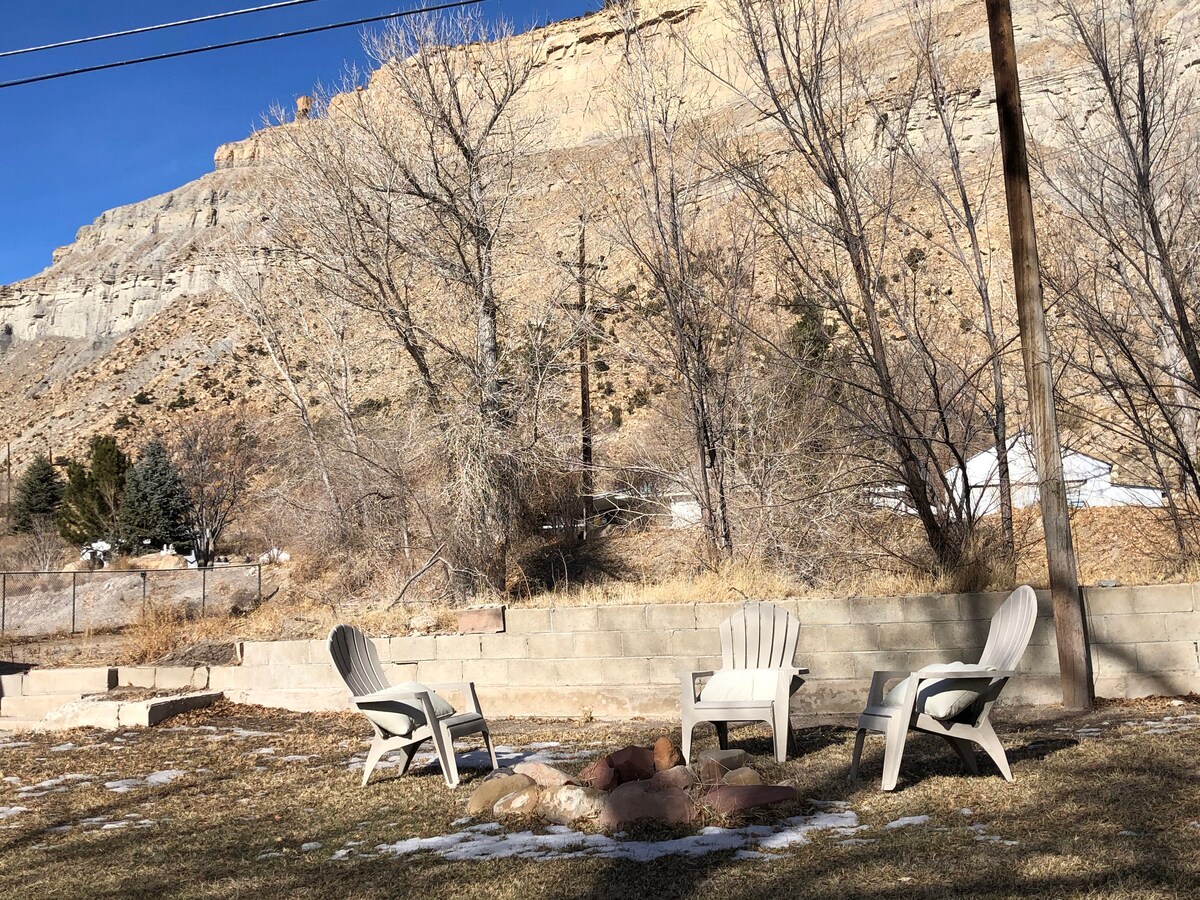 A semi-fenced grass yard is visible, featuring three Adirondack chairs arranged around a stone fire pit. The backdrop includes a rocky terrain with sparse vegetation, lined by tall trees and a distant mountain, under a clear blue sky.