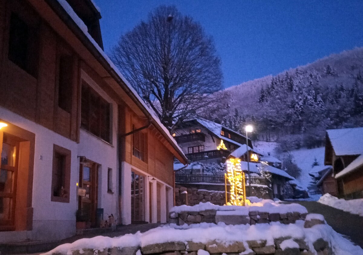 An exterior view of the building is captured during twilight, showcasing warm lighting that highlights architectural features. A snow-covered pathway leads to the entrance, with trees and a hillside adorned in white, creating a serene winter atmosphere.