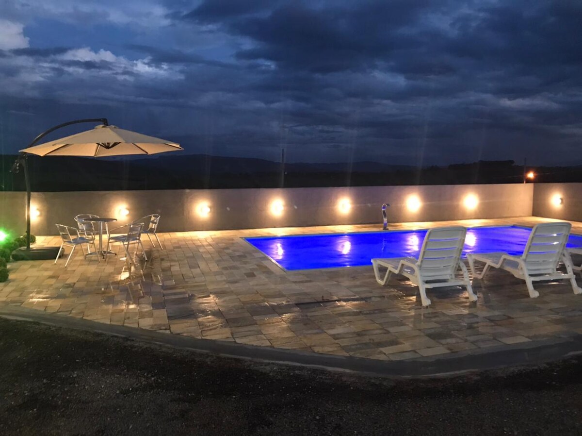 A quiet pool area features a rectangular pool illuminated with soft blue lighting. Surrounding the pool, light-colored patio tiles create a spacious area. Four white lounge chairs and a small table with an umbrella provide seating options, while dim, mounted lights enhance the ambiance.