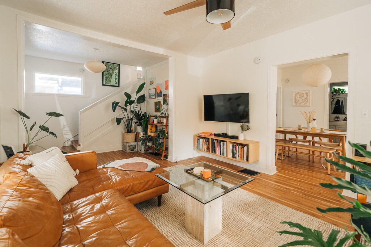 The living area features a curved leather sectional sofa facing a mounted television. A coffee table made of stone and glass sits at the center, surrounded by a natural fiber rug. Plants are placed throughout the space, adding a touch of greenery. A dining area is visible in the background.