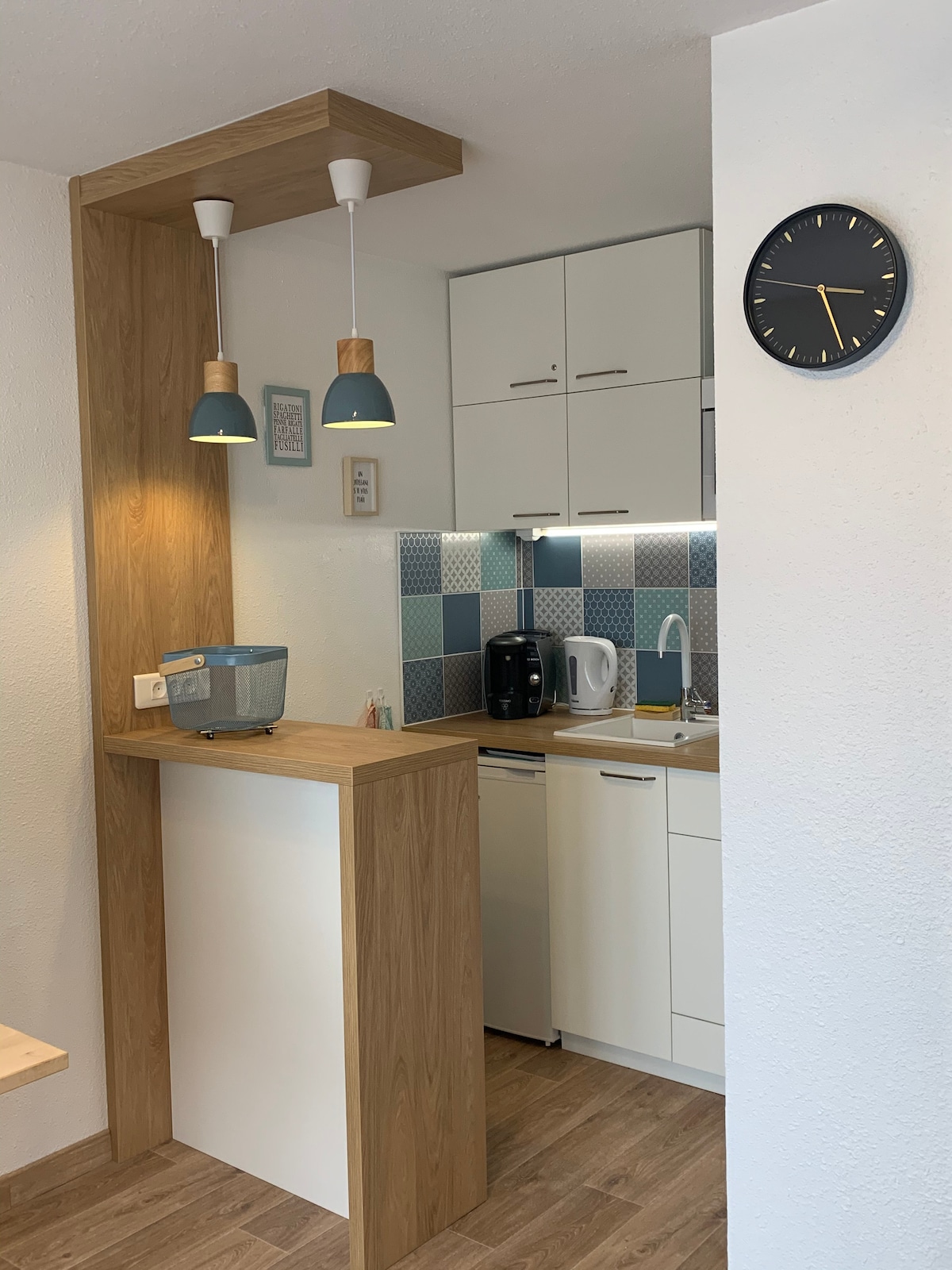 A compact kitchen area is organized with modern cabinetry and a light wood countertop. Hanging light fixtures provide task lighting. Appliances include a coffee machine and kettle, alongside a refrigerator. A wall clock and decorative accents add a practical touch, enhancing the kitchen's functionality.